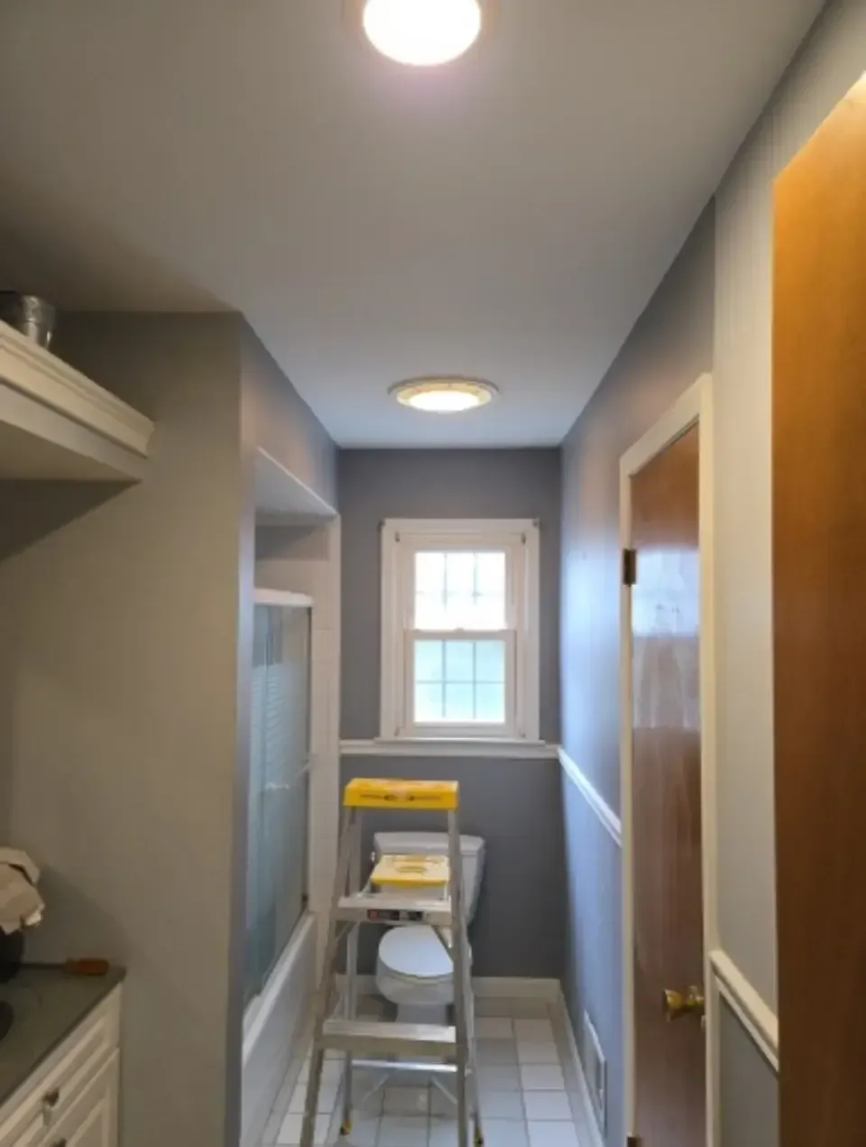 Narrow hallway with gray walls, ceiling lights, a window, and a ladder in the center.