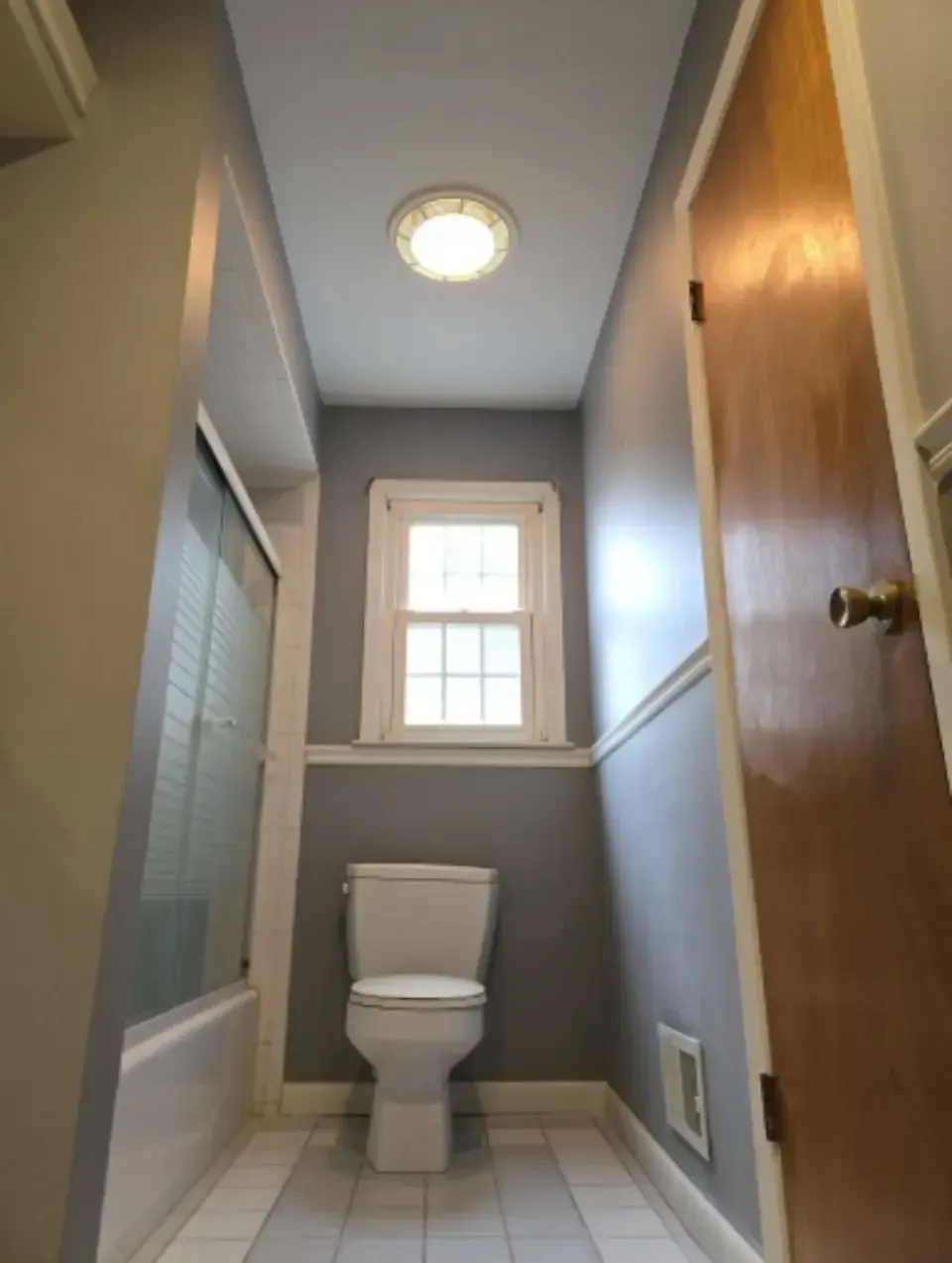 Narrow bathroom hallway with a toilet under a window, gray walls, and a wooden door
