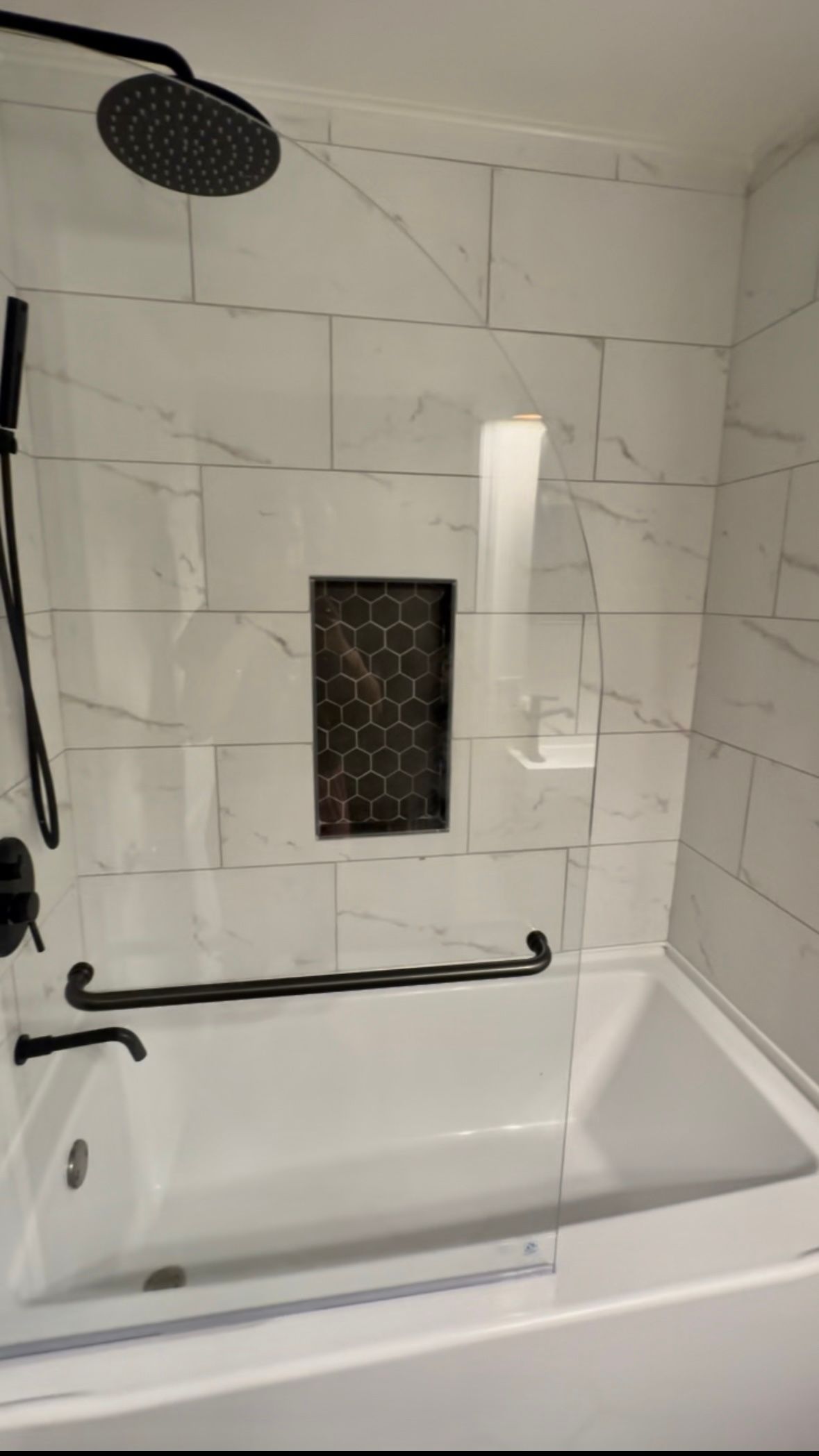 White tiled shower-tub with black fixtures, bench, and a small recessed shelf.