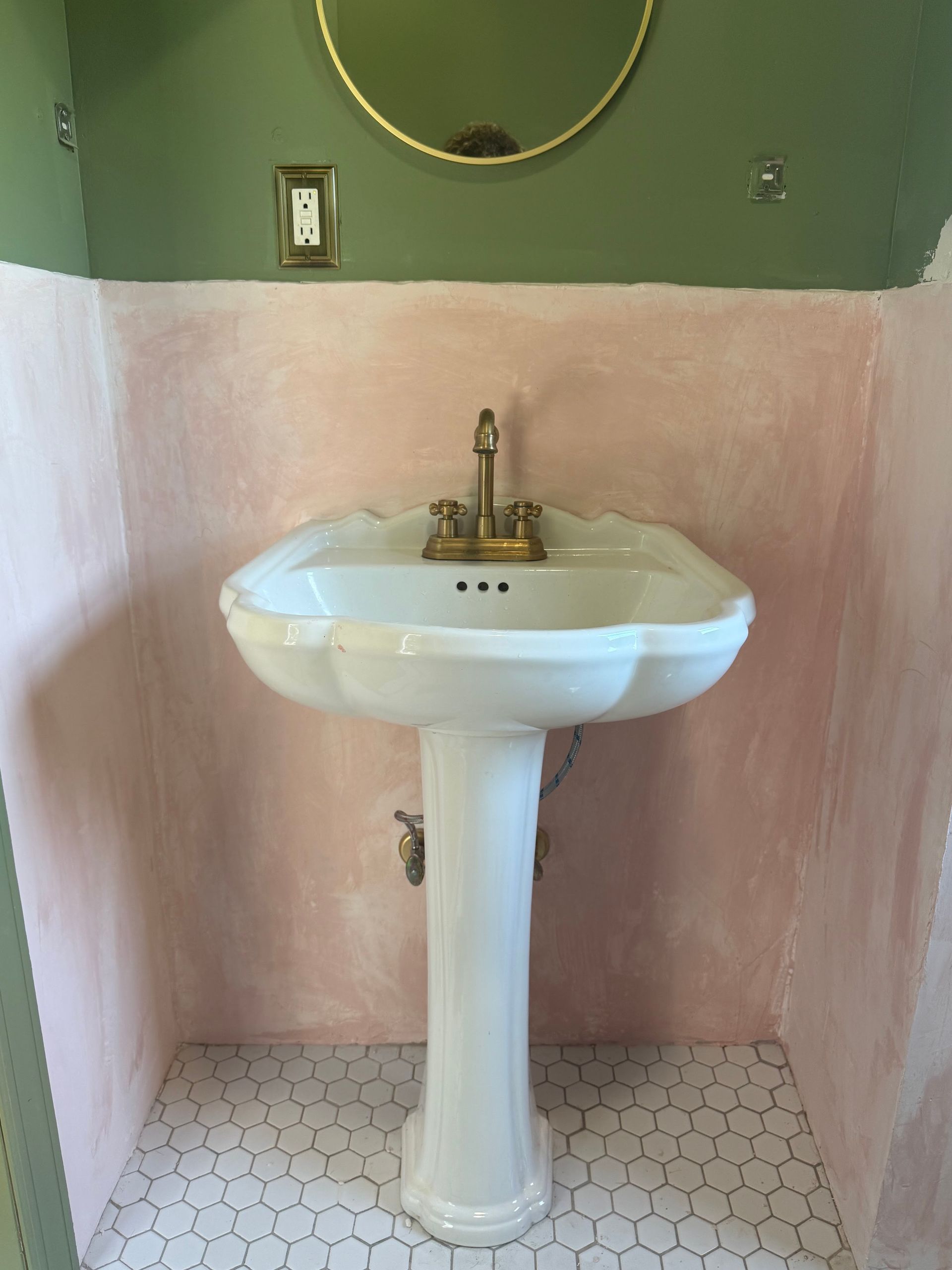 White pedestal sink against a pink-and-green bathroom wall with a round mirror above.
