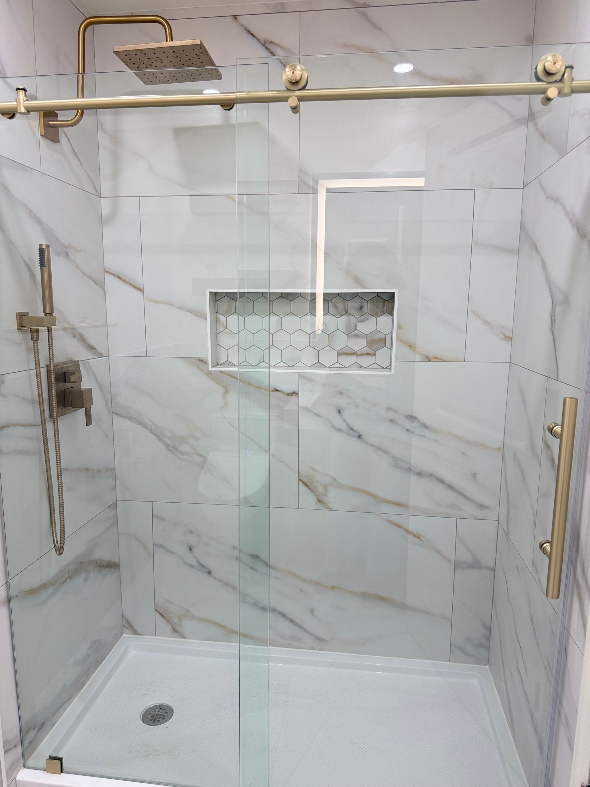 Modern tiled shower with glass door, brass fixtures, white bench, and built-in shelf.