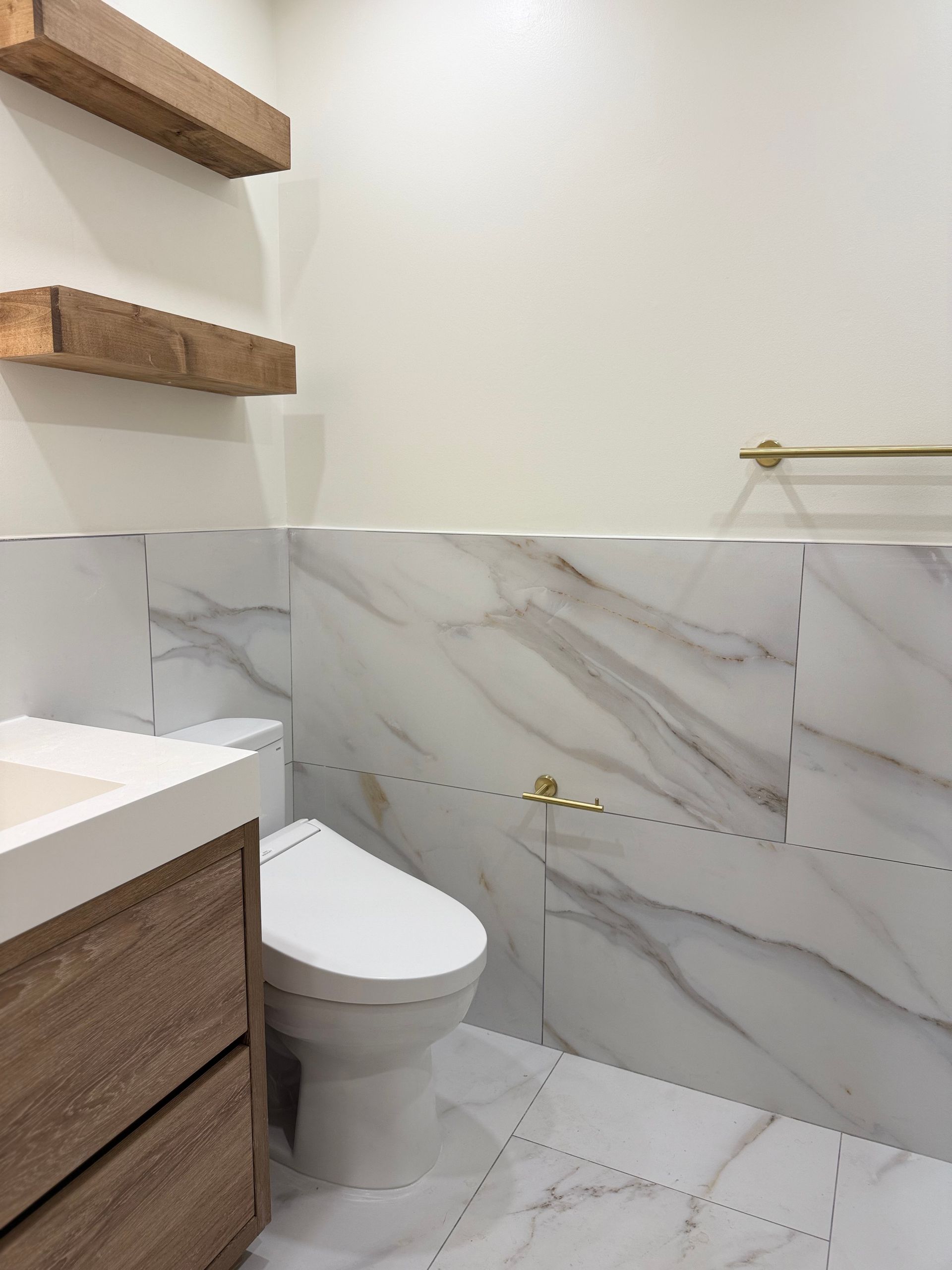 Modern bathroom with white marble-look tile, wooden vanity, toilet, and wall shelves