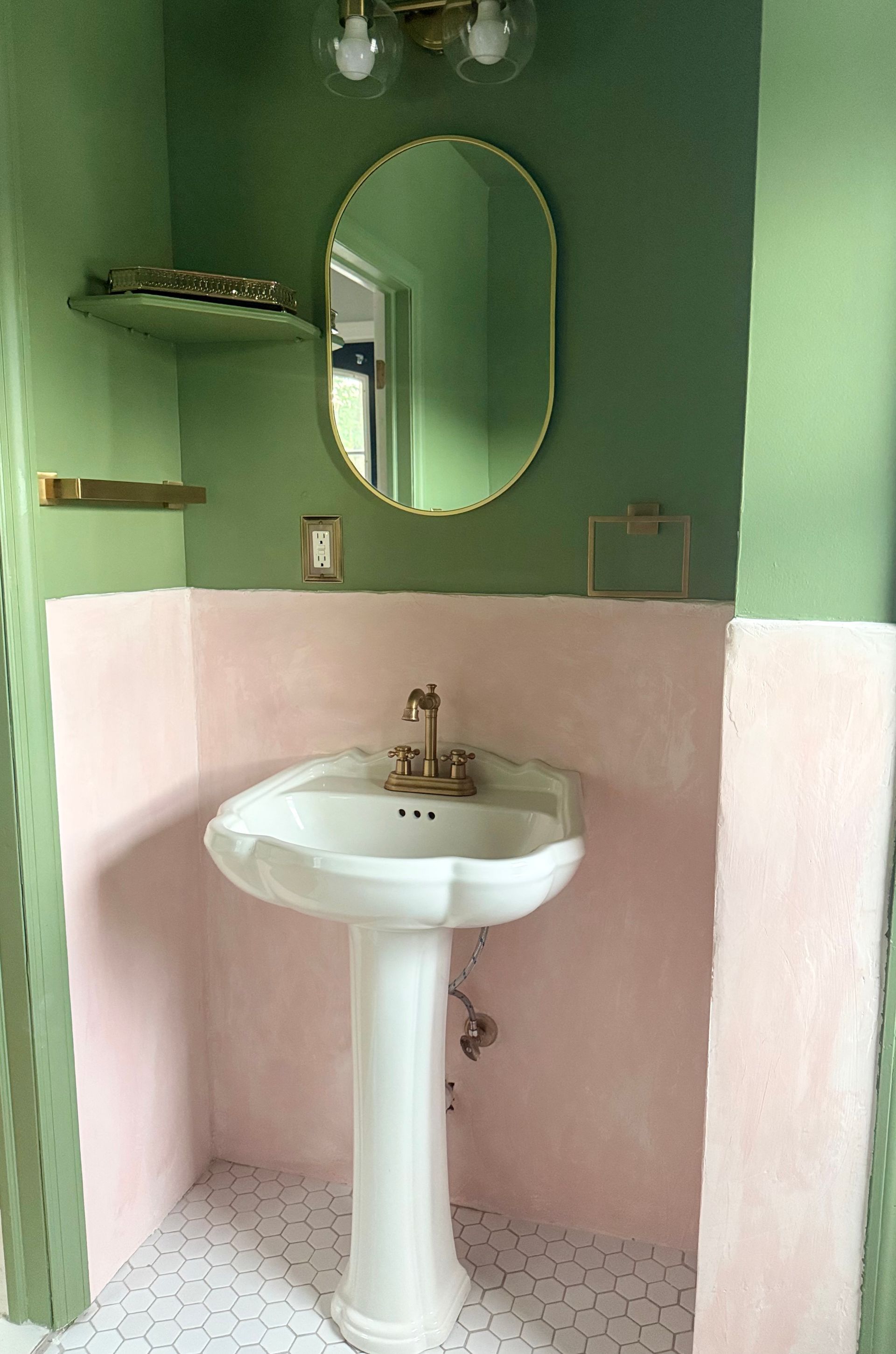 Small bathroom with green and pink walls, white pedestal sink, oval mirror, and wall shelves.