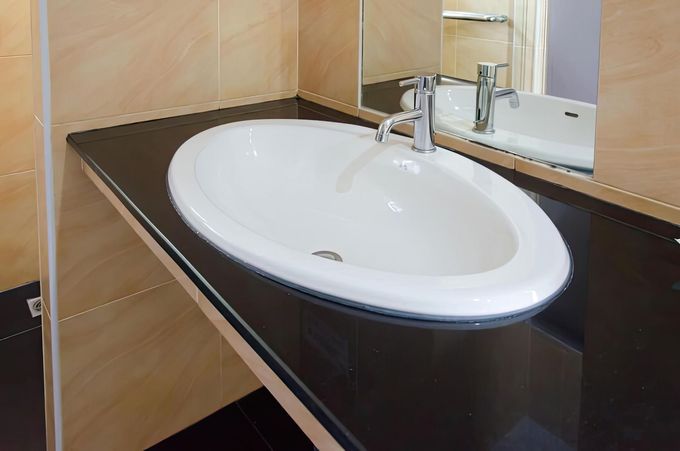 Oval white sink on a dark countertop in a beige-tiled bathroom with a mirror above