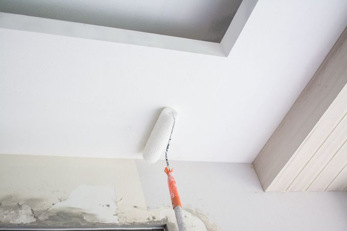 A person painting a white ceiling corner with a paint roller on an extension pole.