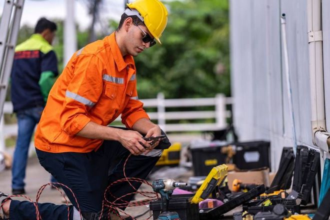 Construction worker kneels, examining tools, wearing orange workwear and a yellow hardhat.