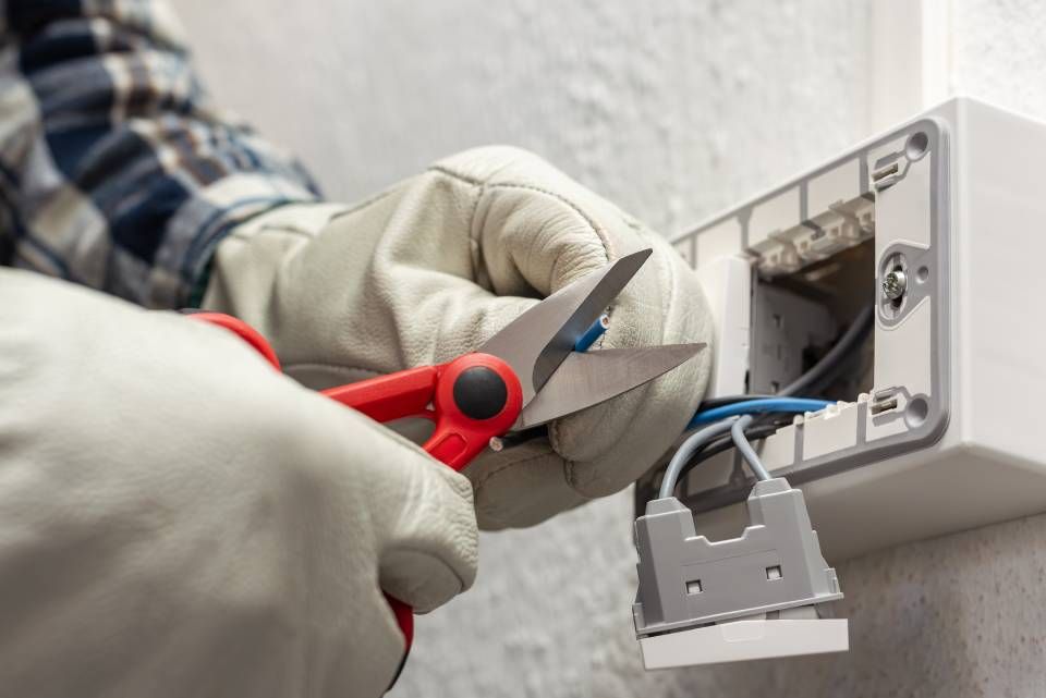 Electrician using scissors to cut wires near a wall outlet, wearing gloves.