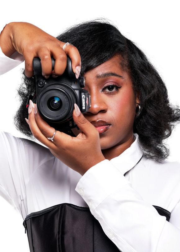 Londyn in white shirt and black corset holding a camera up to their eye against a plain background