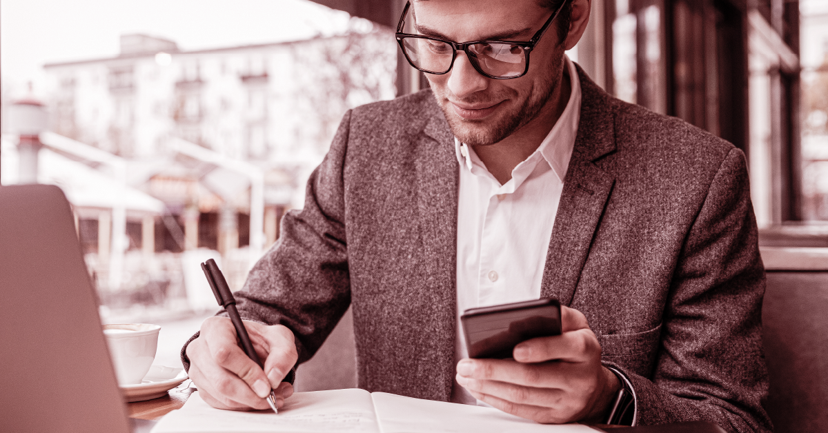 Profissional fazendo anotações com caneta e celular ao lado de notebook e café em ambiente externo.