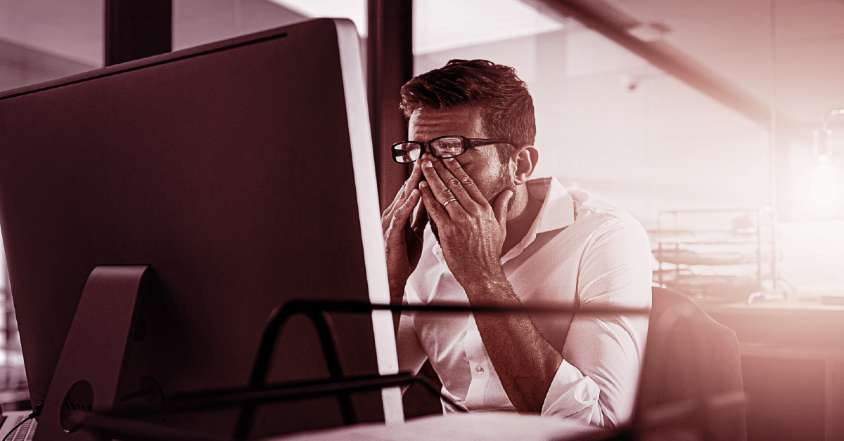 Profissional estressado com as mãos no rosto em frente ao computador no ambiente de trabalho.