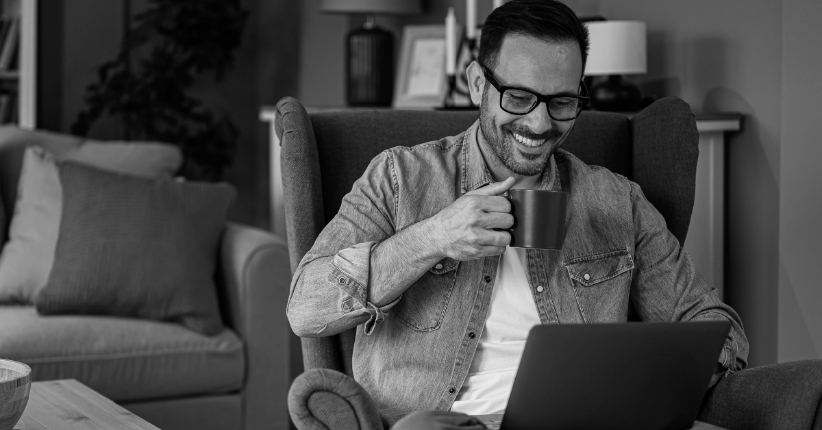 Homem usando laptop, sorrindo, segurando uma caneca. Imagem em preto e branco.