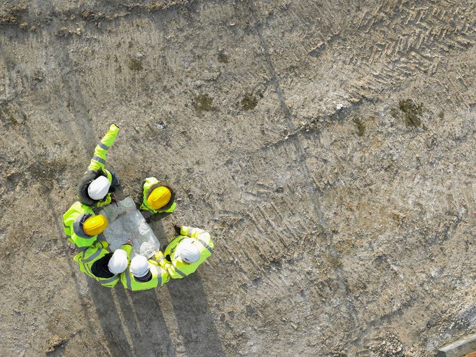 Operai edili con giubbotti gialli e caschi bianchi si accalcano attorno a un blocco di cemento in un cantiere edile.