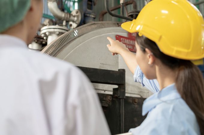 Una donna con un casco giallo indica un'attrezzatura industriale, insieme a un'altra persona con un camice bianco.