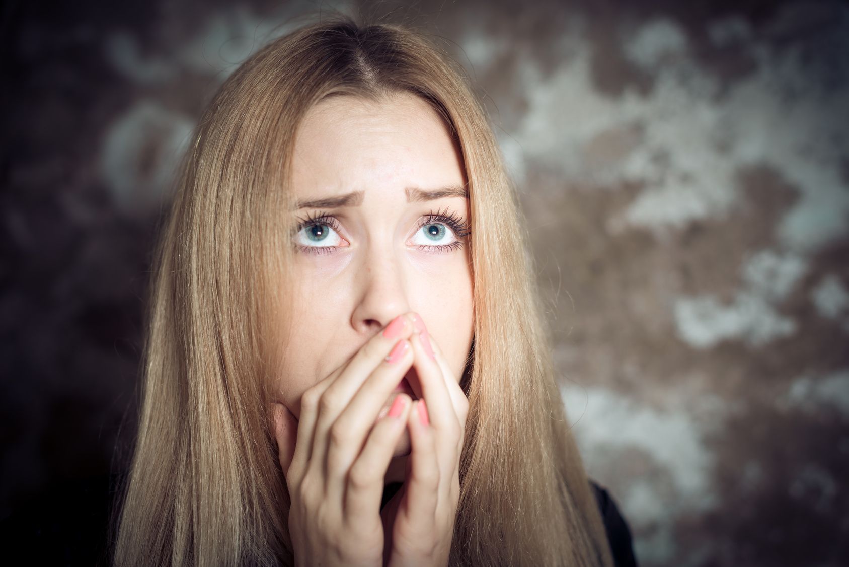 Donna con lunghi capelli biondi, che guarda in alto con un'espressione angosciata, con le mani che coprono la bocca.
