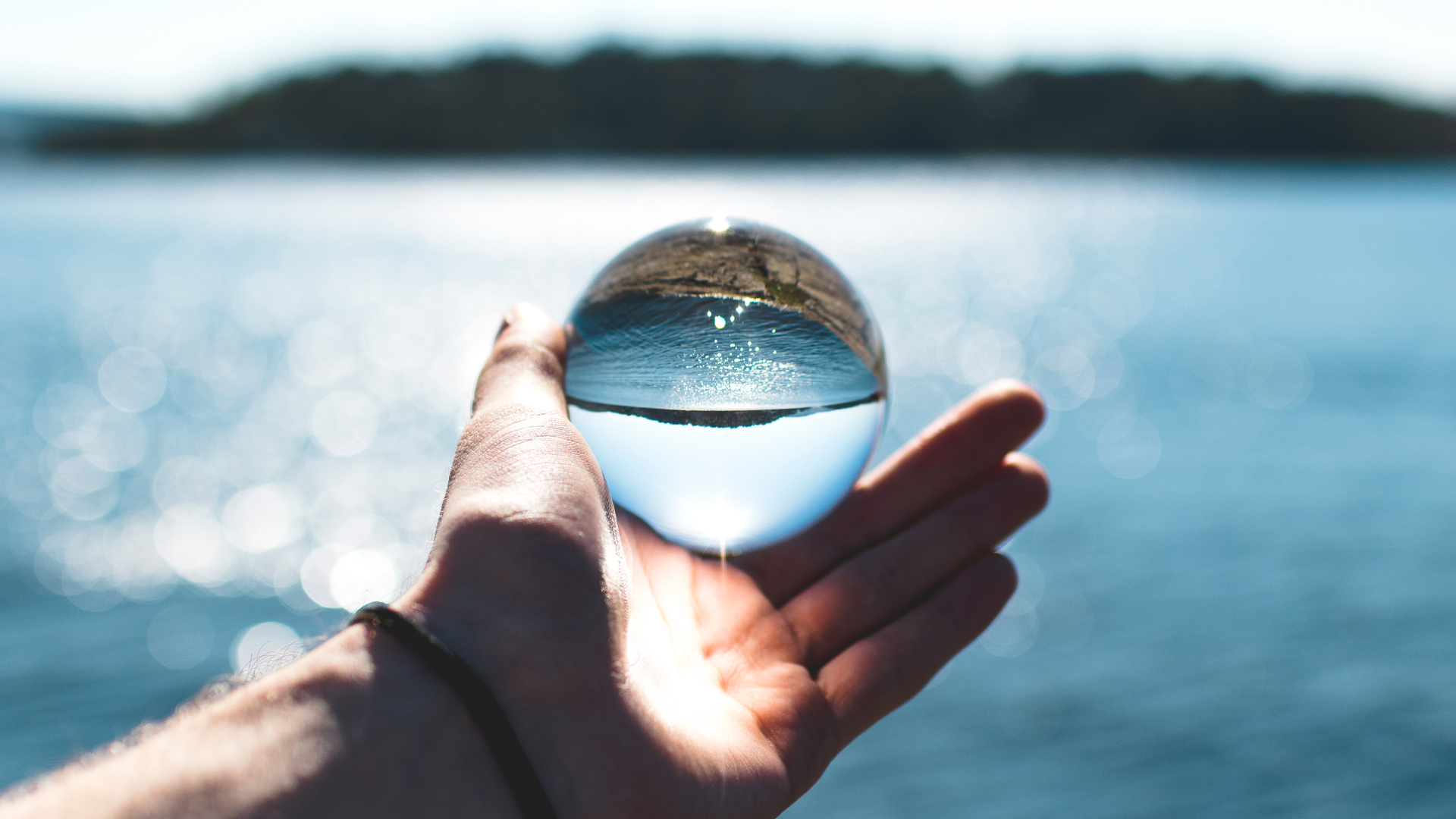 Mano che tiene una sfera di vetro trasparente che riflette un paesaggio marino; acqua blu, giornata di sole.