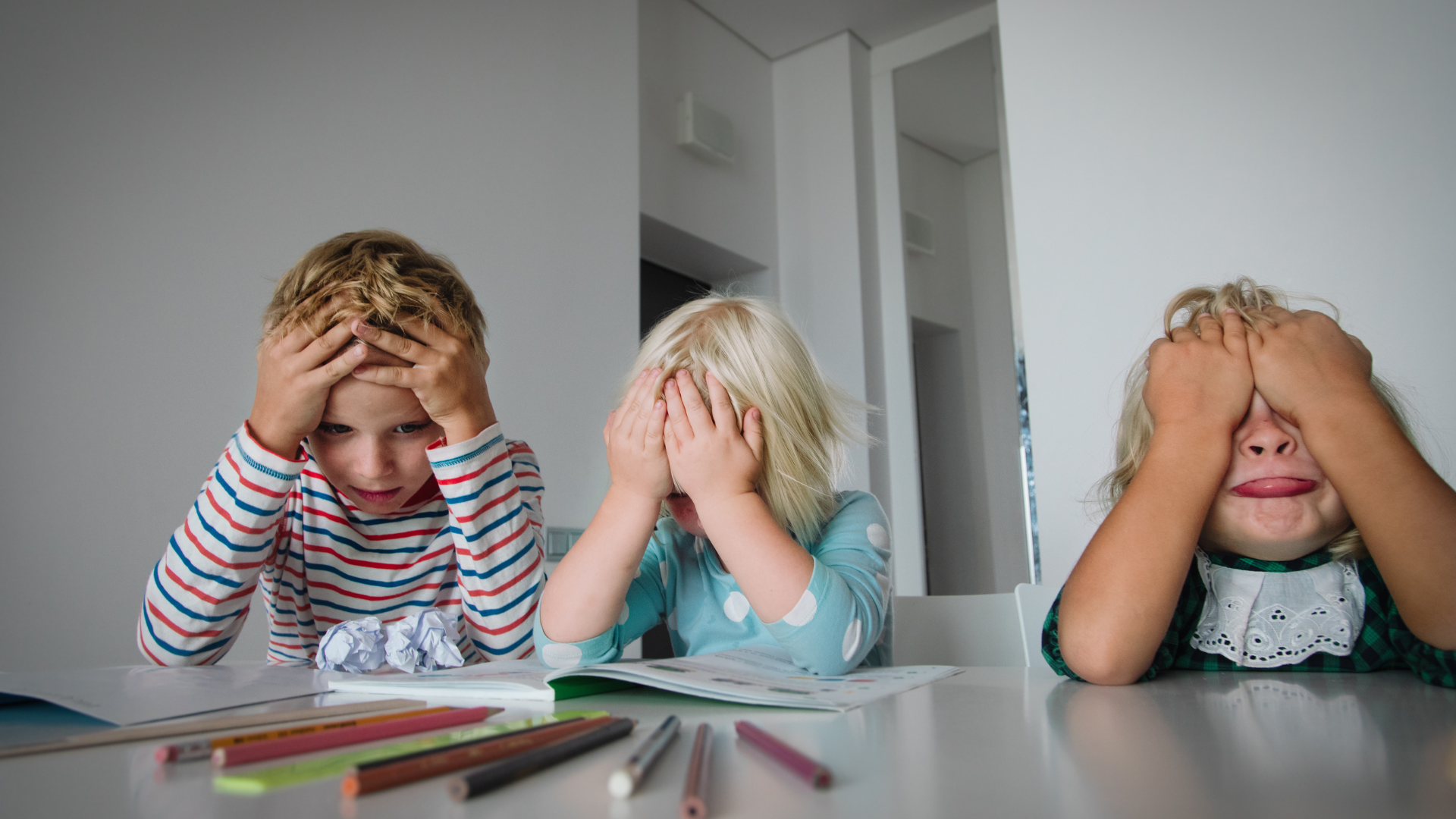 Tre bambini con le mani sul viso, con aria frustrata, seduti a un tavolo con i colori per colorare.