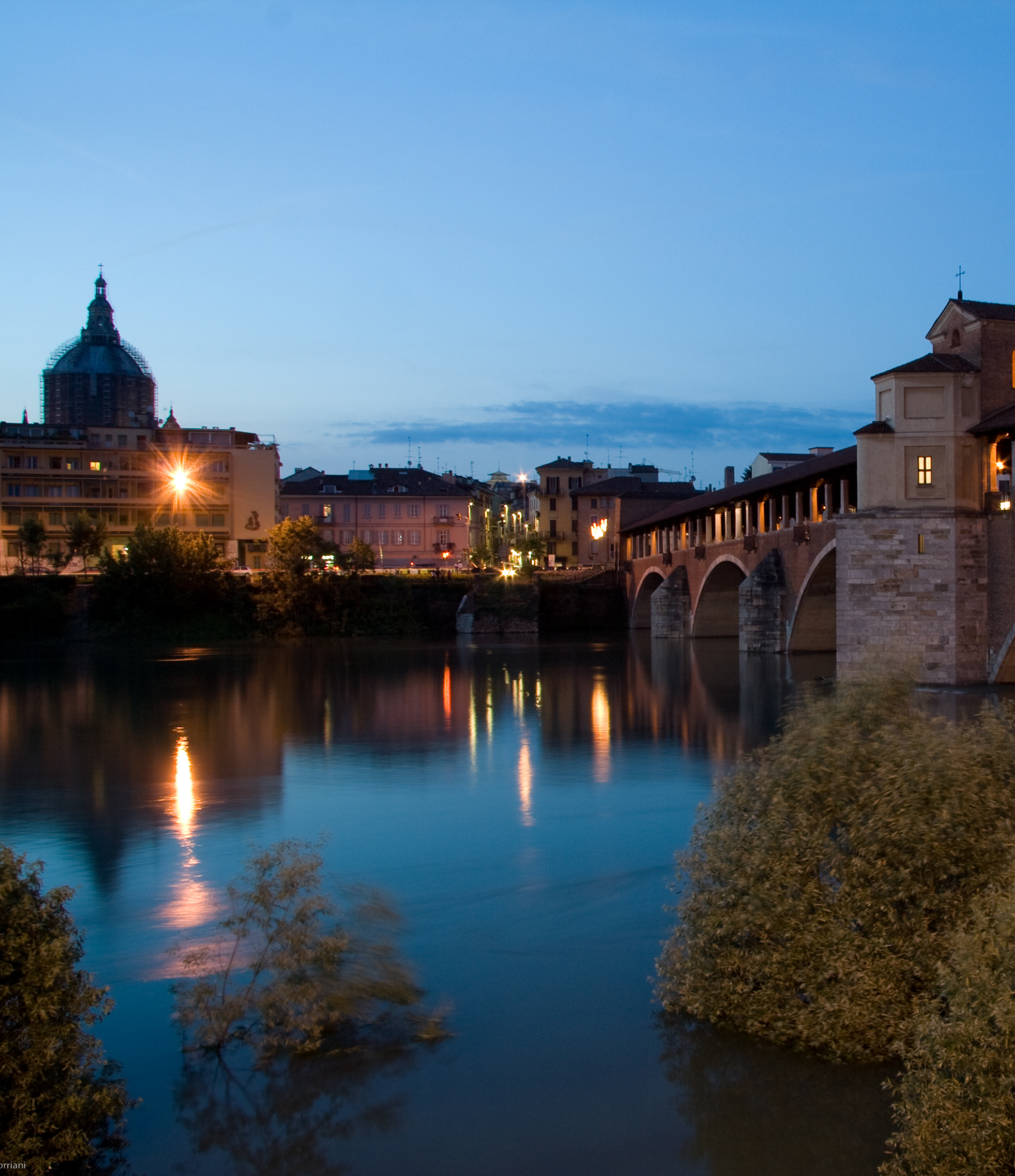 Vista notturna di Pavia, Italia, con il Ponte Coperto sul fiume, edifici e una chiesa con cupola sotto un cielo azzurro.