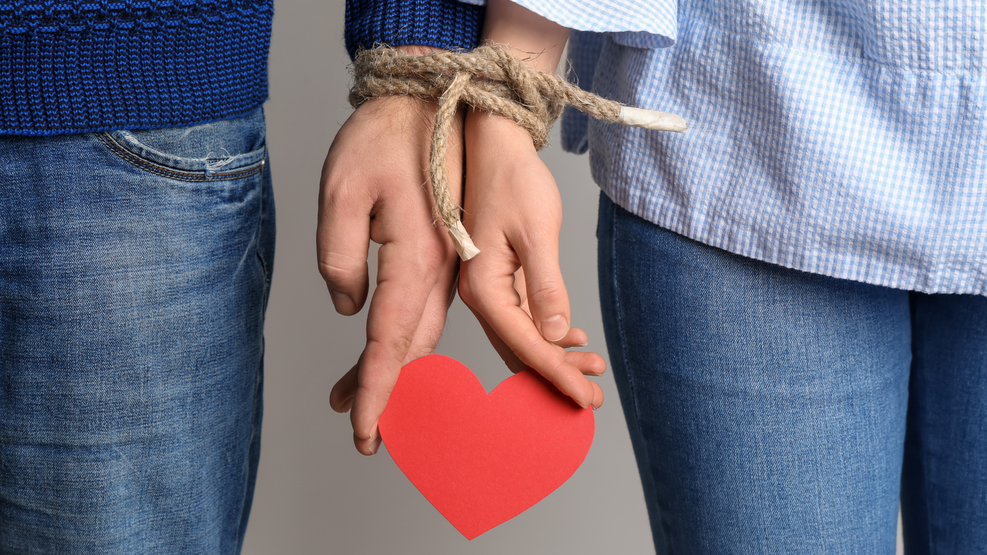 Due mani legate da una corda che tengono un cuore rosso. Una persona indossa una camicia blu e una con un maglione blu.