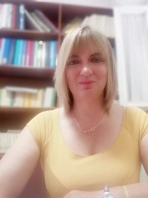 Donna con i capelli biondi che indossa una maglietta gialla, sorridente, davanti a una libreria.