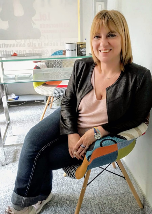 Donna seduta su una sedia colorata, con indosso una giacca nera e jeans, sorridente in un ambiente d'ufficio.