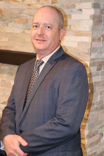 Timothy Bell in a gray suit standing beside a stone wall, smiling slightly.