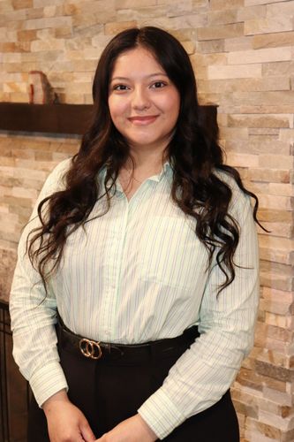 Melany Perez standing indoors against a stone wall, wearing a white blouse and black skirt, smiling at the camera.