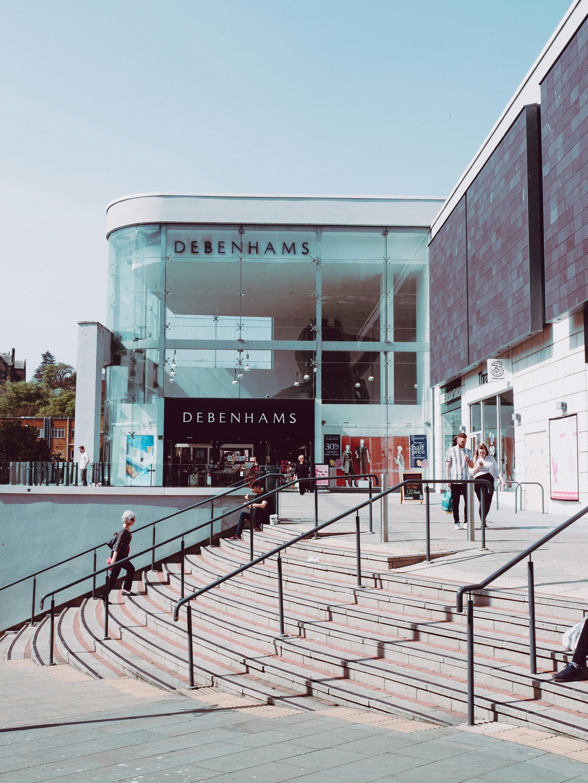 Entrance to a Debenhams store with glass front, steps leading up, and people walking.