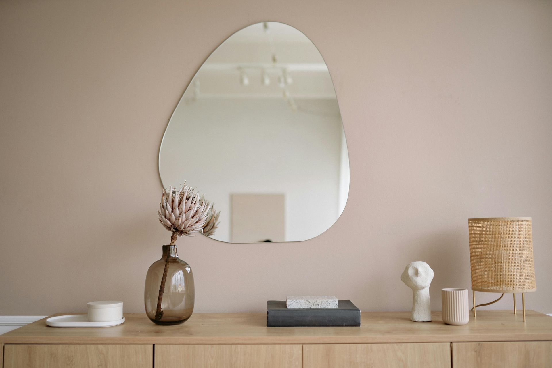 Mirror on a beige wall above a wooden console, featuring decor like a lamp, vase, and books.