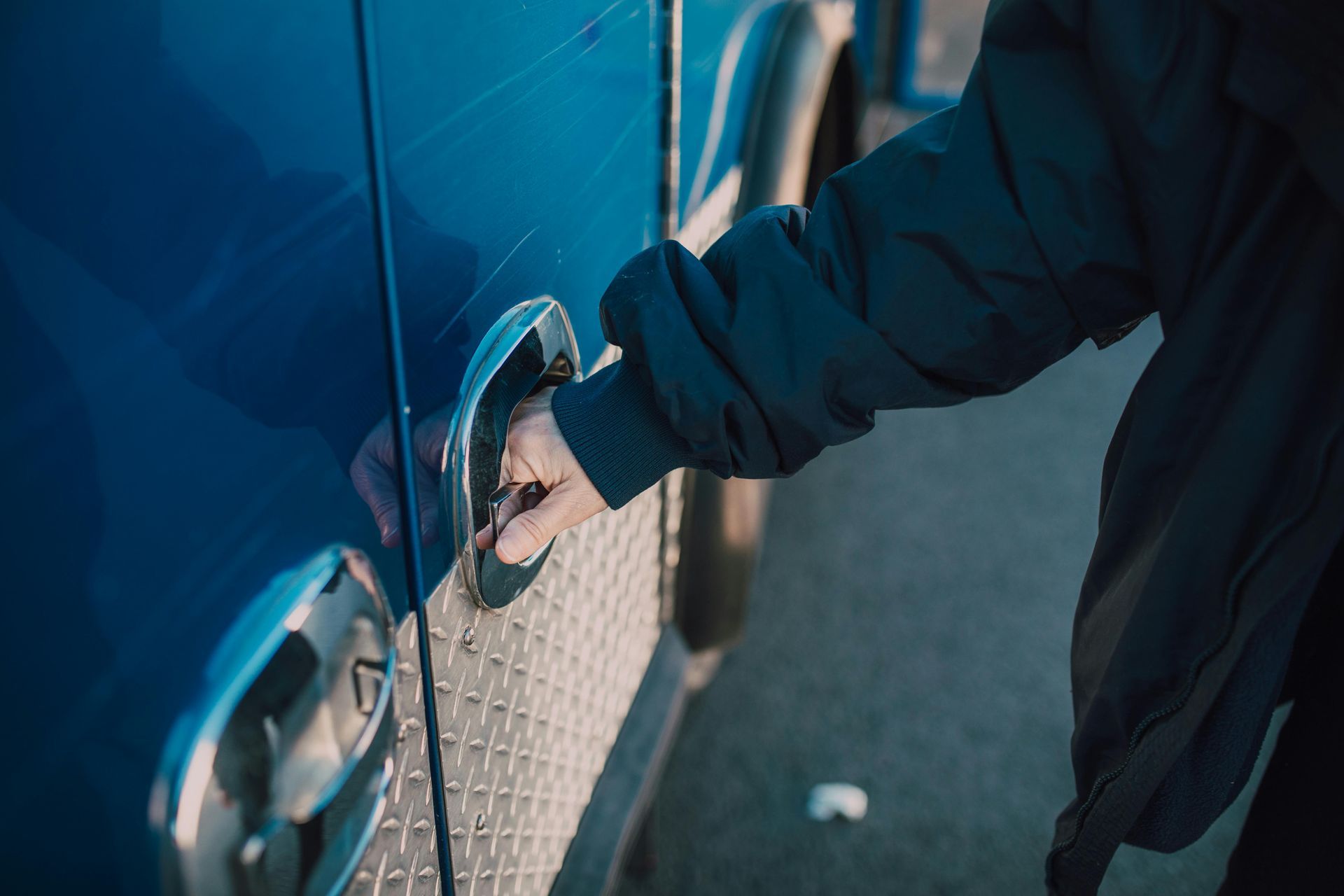 Person's hand pulling the handle of a blue vehicle door. Black jacket sleeve visible.