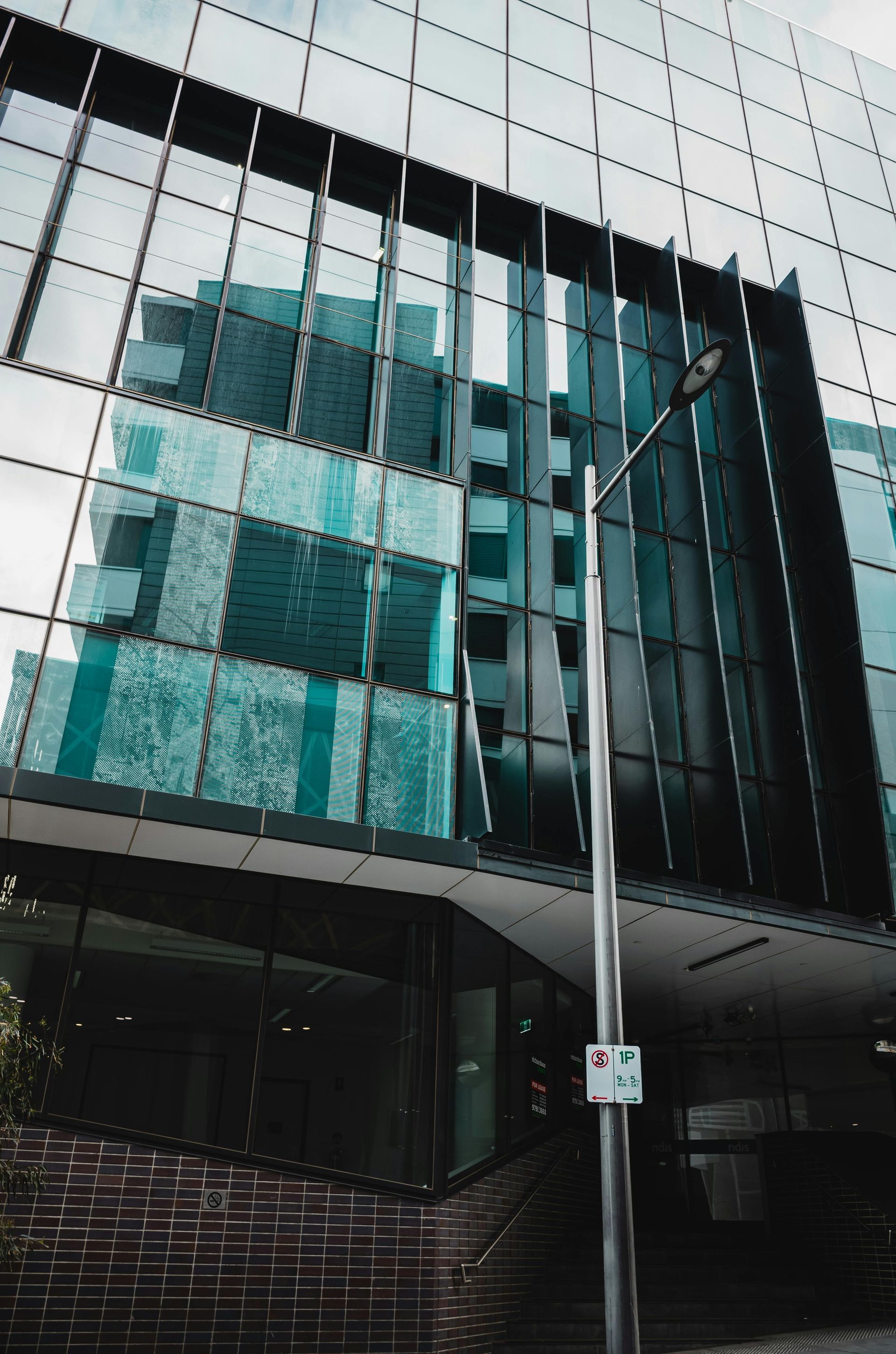 Modern glass building with reflections of teal buildings and a street lamp.