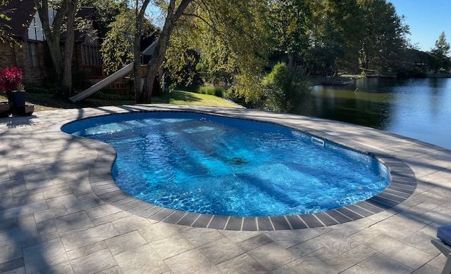 Pool with blue water and surrounding stone patio next to a lake.