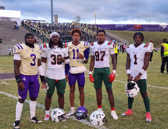 Five football players posing on a field in white jerseys and helmets, with a stadium crowd behind them.