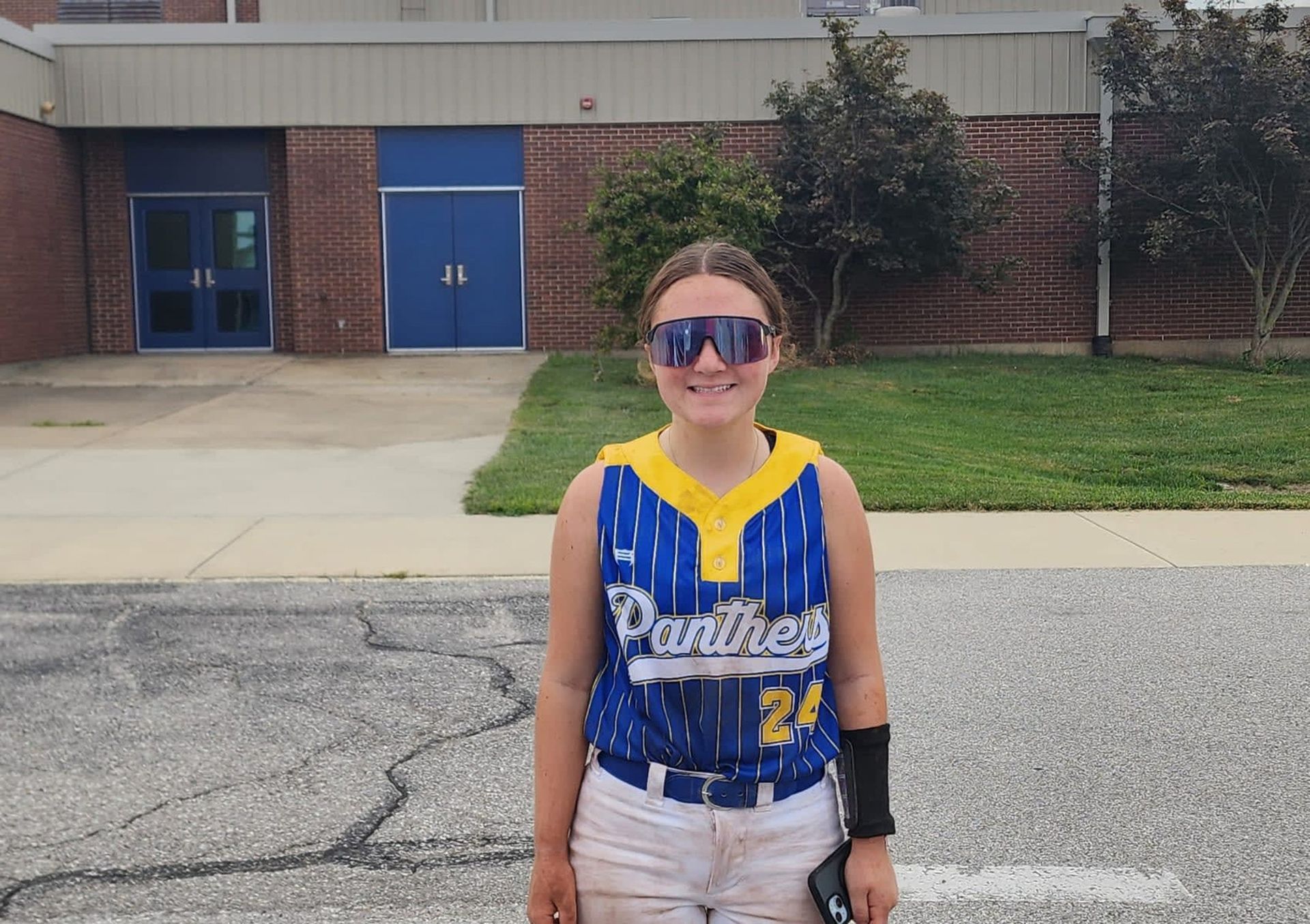 Person in a blue-and-yellow sports jersey and sunglasses standing outside a brick building.