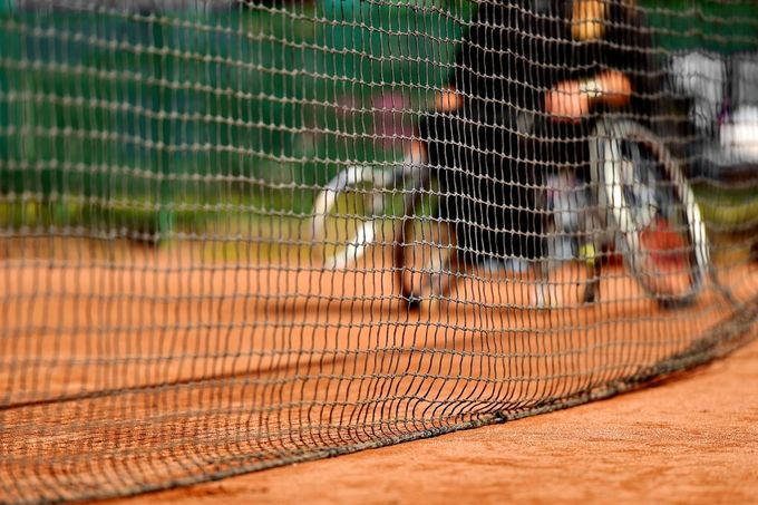 Tennis player on clay court behind net, with green fence in the background