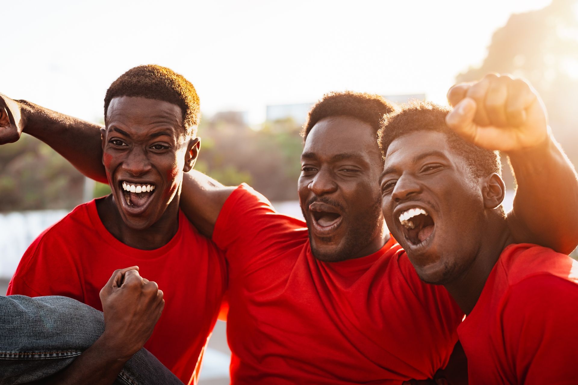 Three friends in red shirts cheering and smiling outdoors in warm sunlight