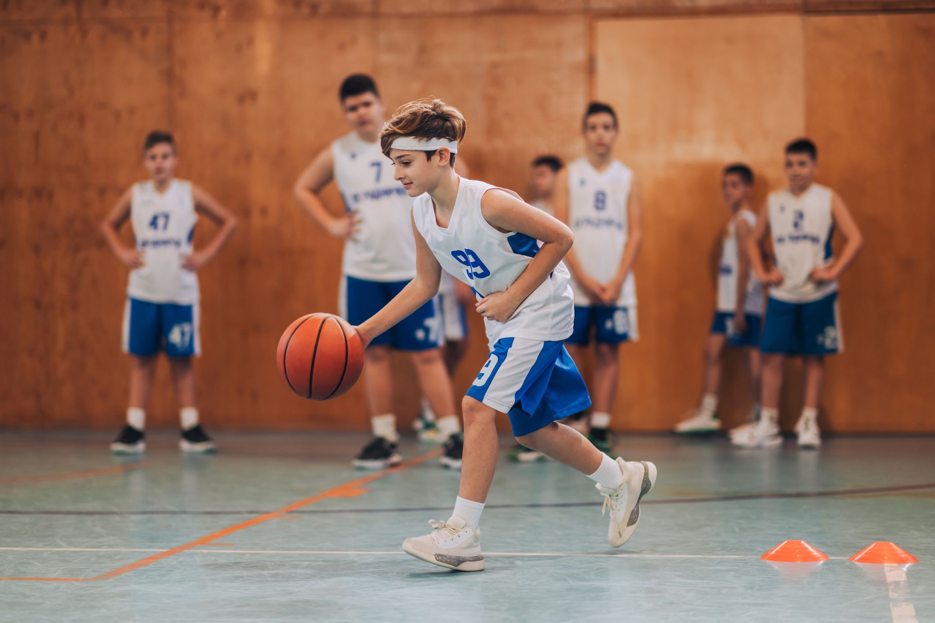 Basketball player dribbling in a gym during practice, with teammates and cones in the background.