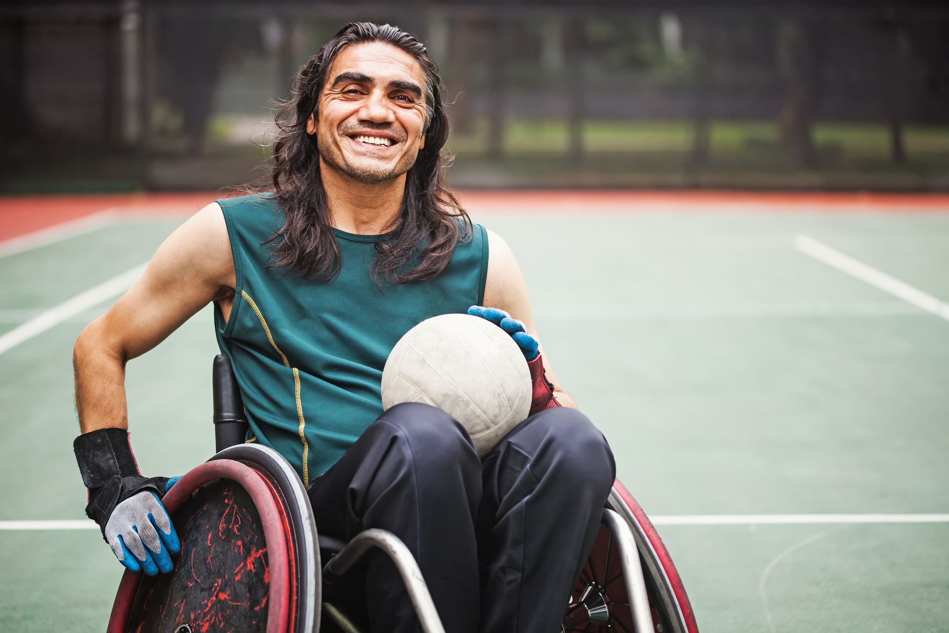 Wheelchair tennis player smiling on a court, holding a ball in his lap.