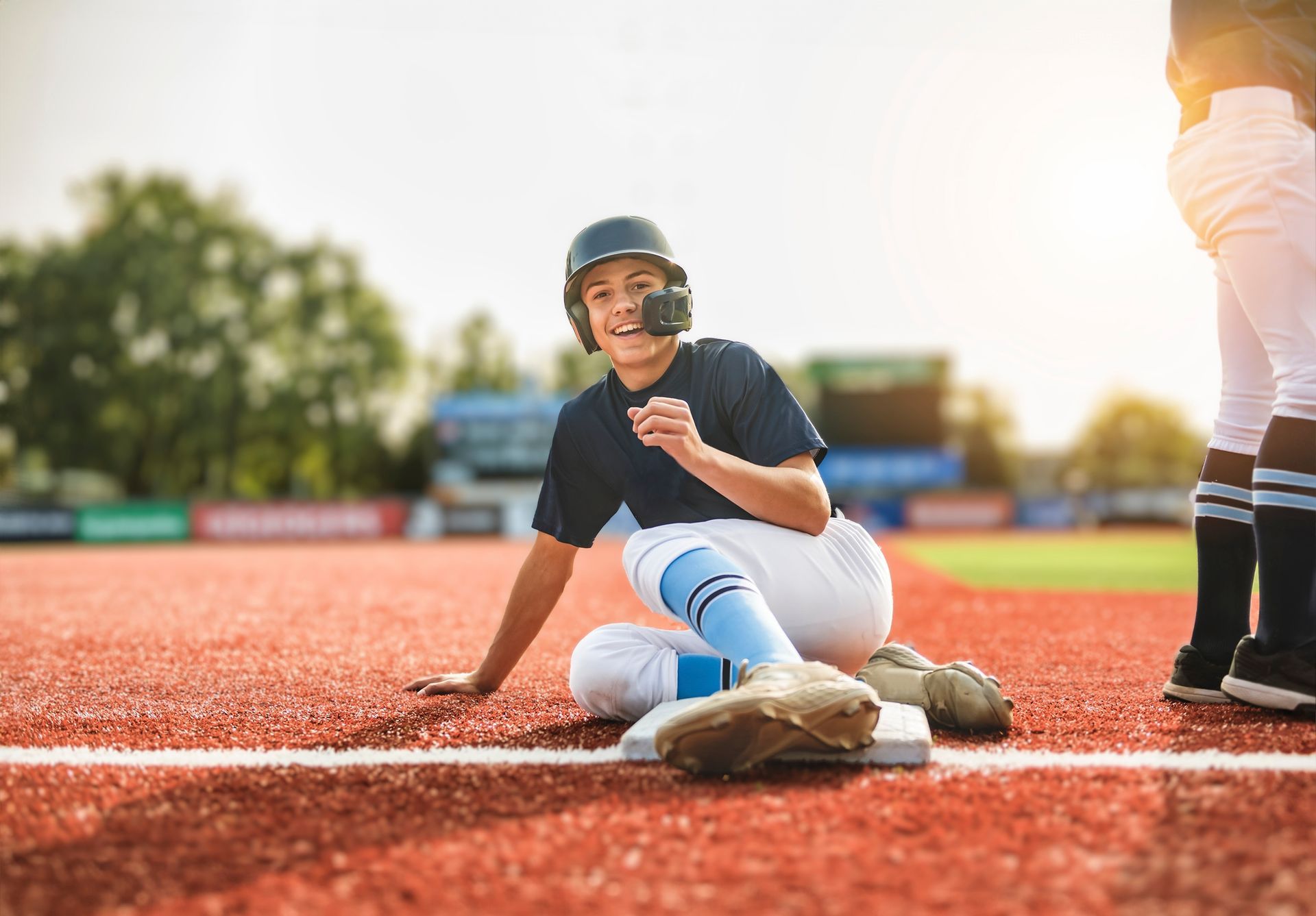 Baseball player sitting on a field, smiling with helmet and batting gear near home plate