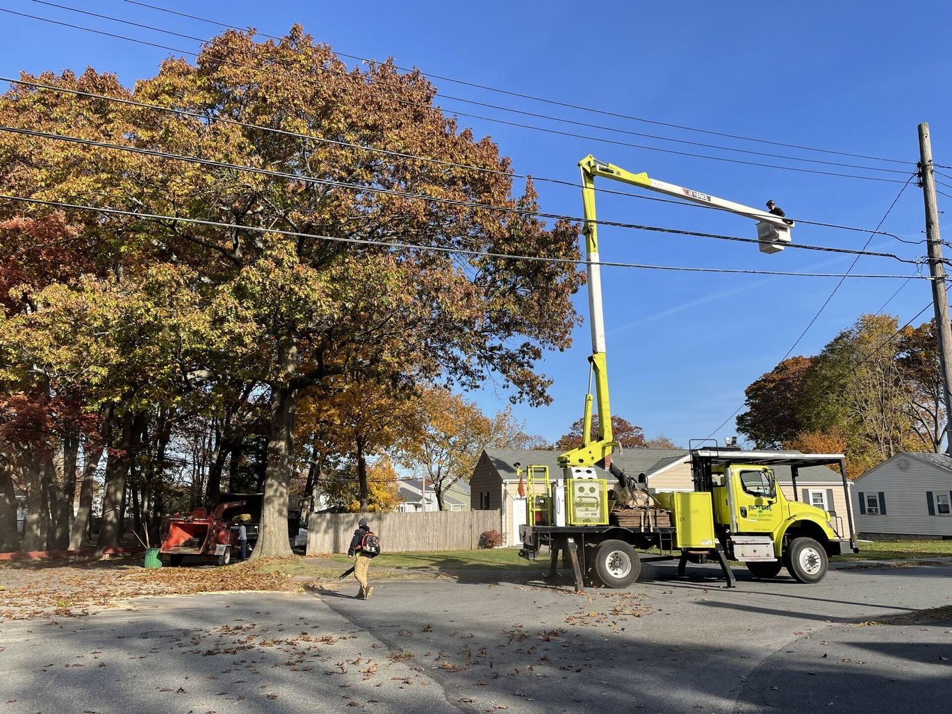 Truck with an elevated bucket trimming tree branches near power lines. Autumn leaves, houses, clear sky.