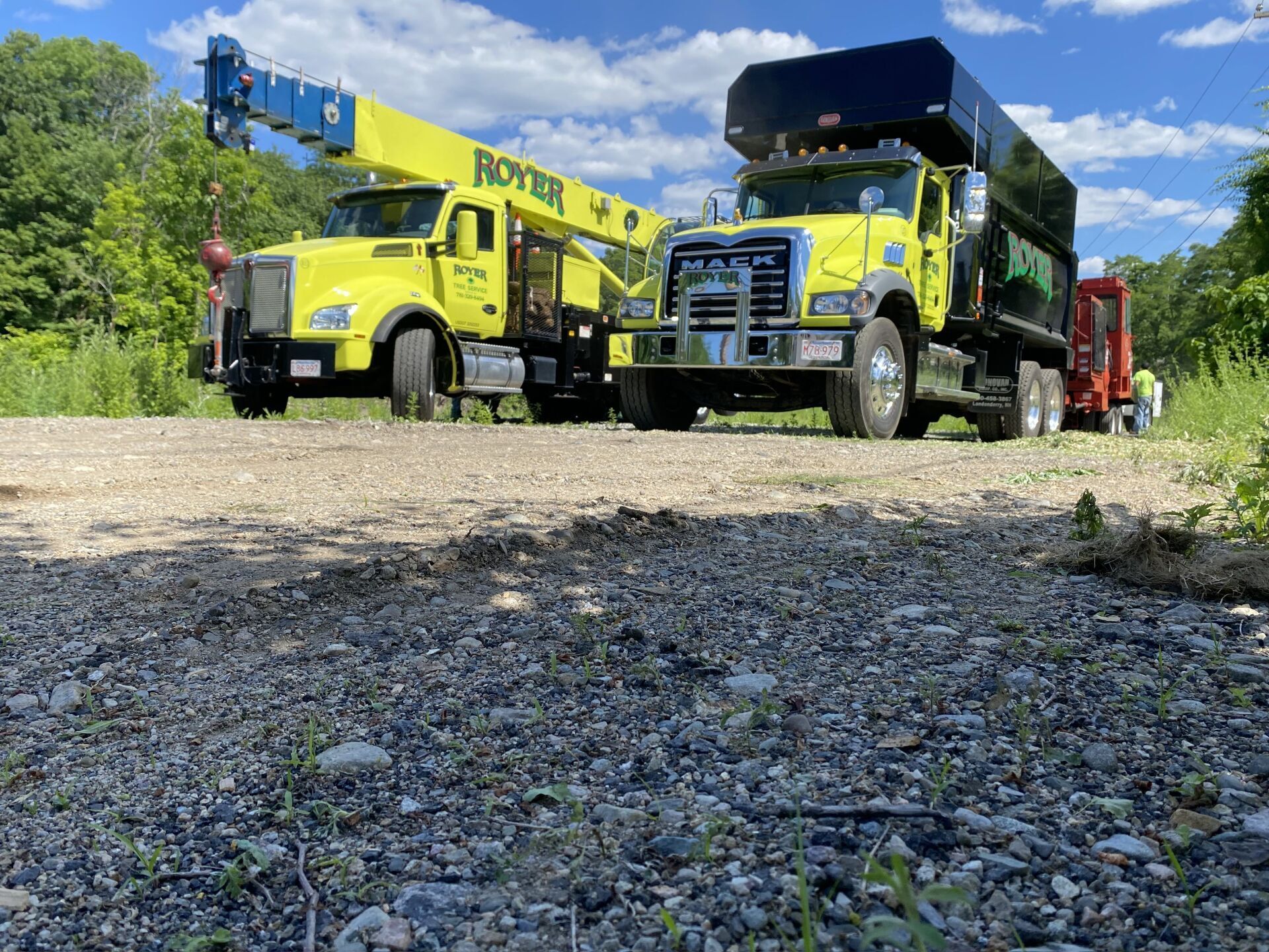 Two yellow work trucks parked on a gravel road, one with a crane, and another a dump truck.