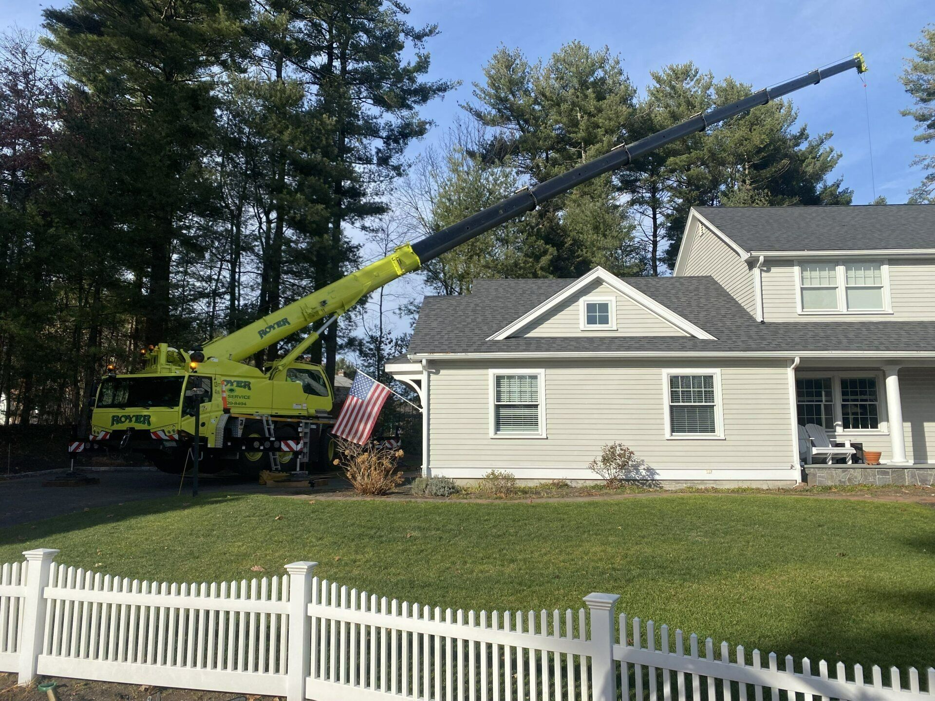 A large yellow crane positioned near a light-colored house with a black roof. A white picket fence is in the foreground.