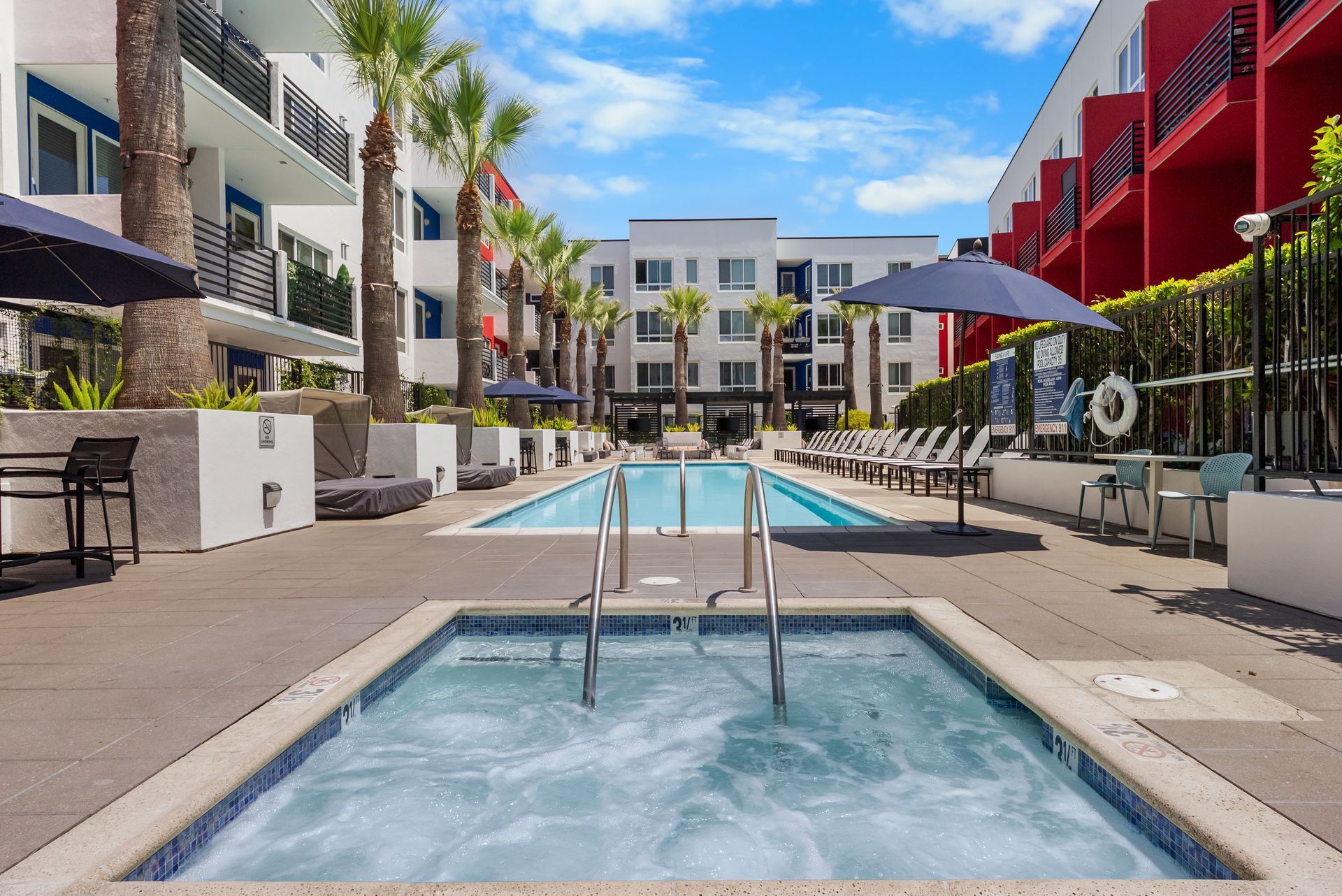 Jacuzzi spa and swimming pool surrounded by lounge chairs and sun umbrellas in building courtyard