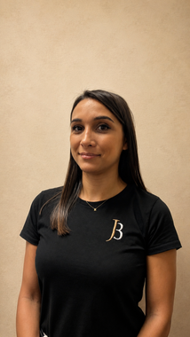 Woman in a black T-shirt with a gold “B” logo, standing against a beige wall.