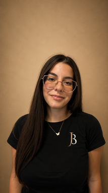 Portrait of a woman with long dark hair and glasses, wearing a black shirt, standing against a tan wall