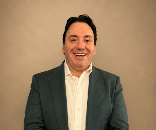 Smiling man in a teal blazer and white shirt against a plain beige background