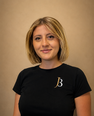 Woman in a black T-shirt with a gold “B” logo, standing against a plain beige background.