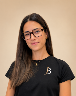 Woman with long dark hair and glasses, wearing a black T-shirt with a white “B,” against a beige background