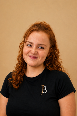 Smiling woman with curly red hair wearing a black shirt against a beige wall