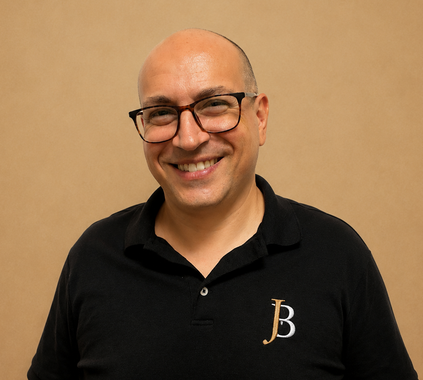 Smiling bald man wearing glasses and a black polo shirt against a tan background
