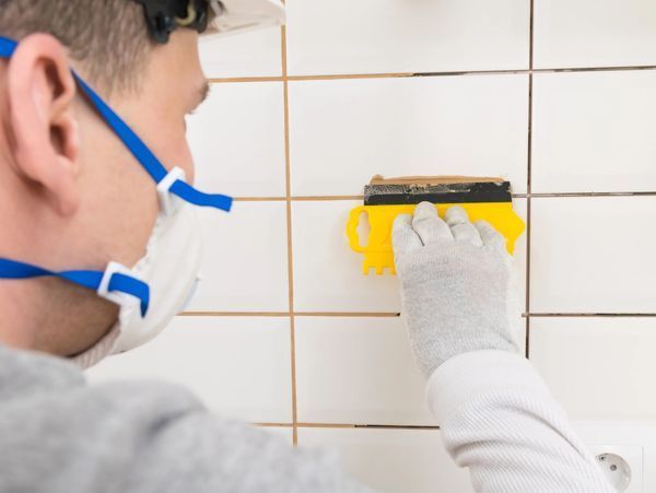 Person applying yellow grout to white tiled wall with a trowel while wearing a mask and gloves